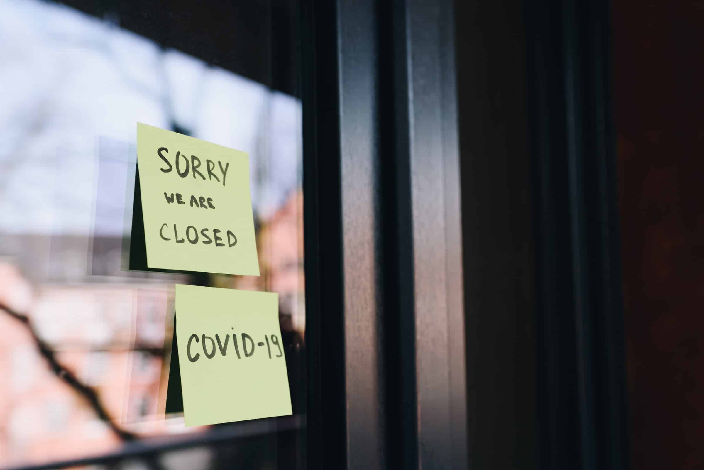 A photo of two Post-it notes stuck on a business's door that read (top to bottom) "Sorry We Are Closed" and "COVID-19".
