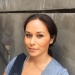 Nina Dewi smiles close-lipped at the camera, posing against a gray wall with her hair back in a ponytail.