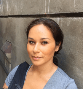 Nina Dewi smiles close-lipped at the camera, posing against a gray wall with her hair back in a ponytail.