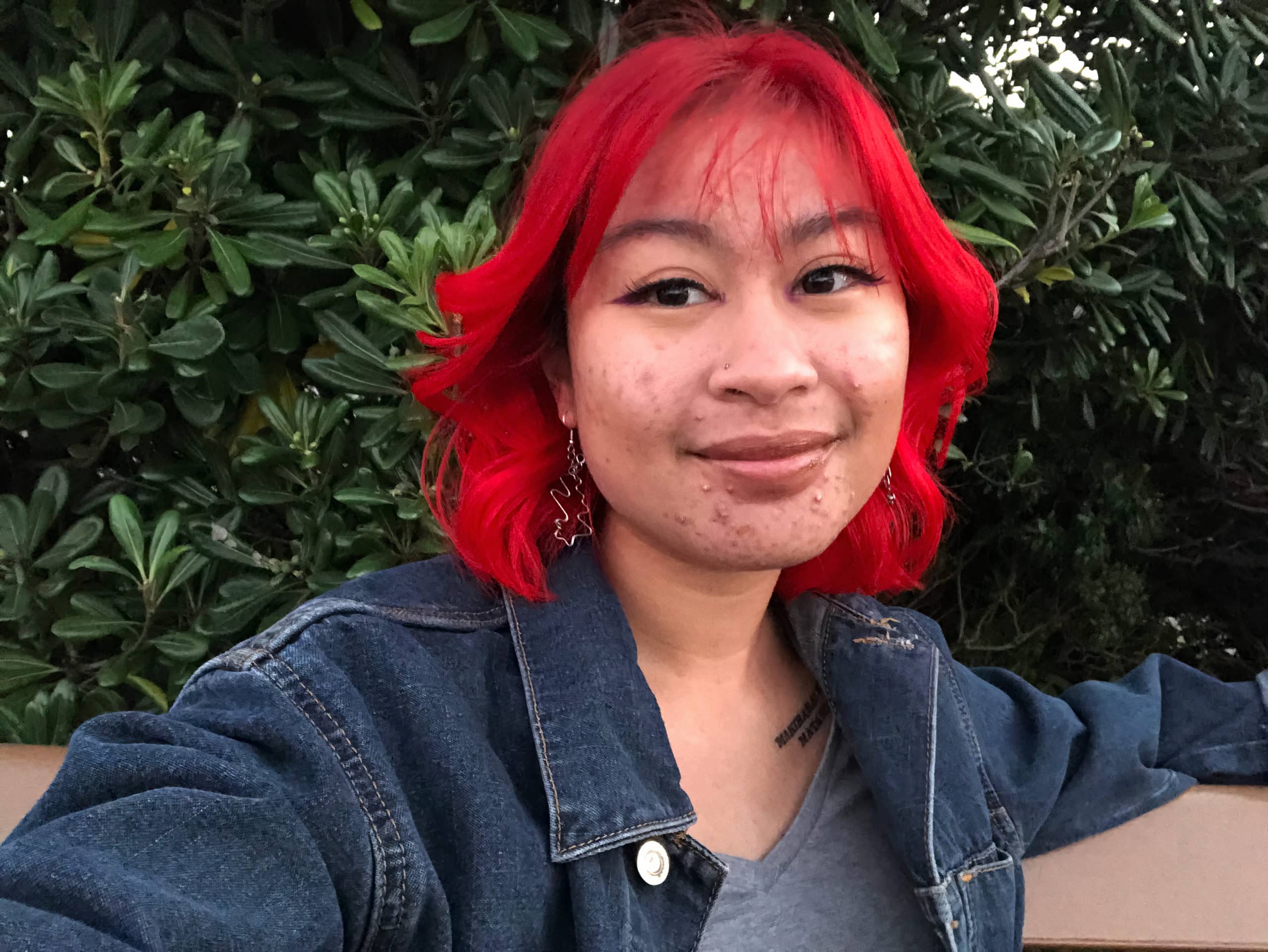 Janelle Salanga, The Objective’s deputy editor, smiles at the camera in front of a bush. They’re wearing a denim jacket.