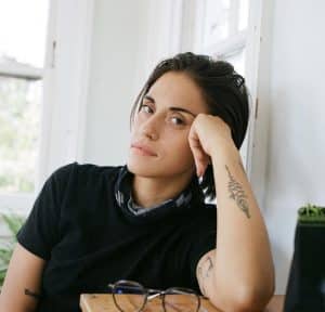 Liana DeMasi props their face up with their elbow, looking at the camera seriously.