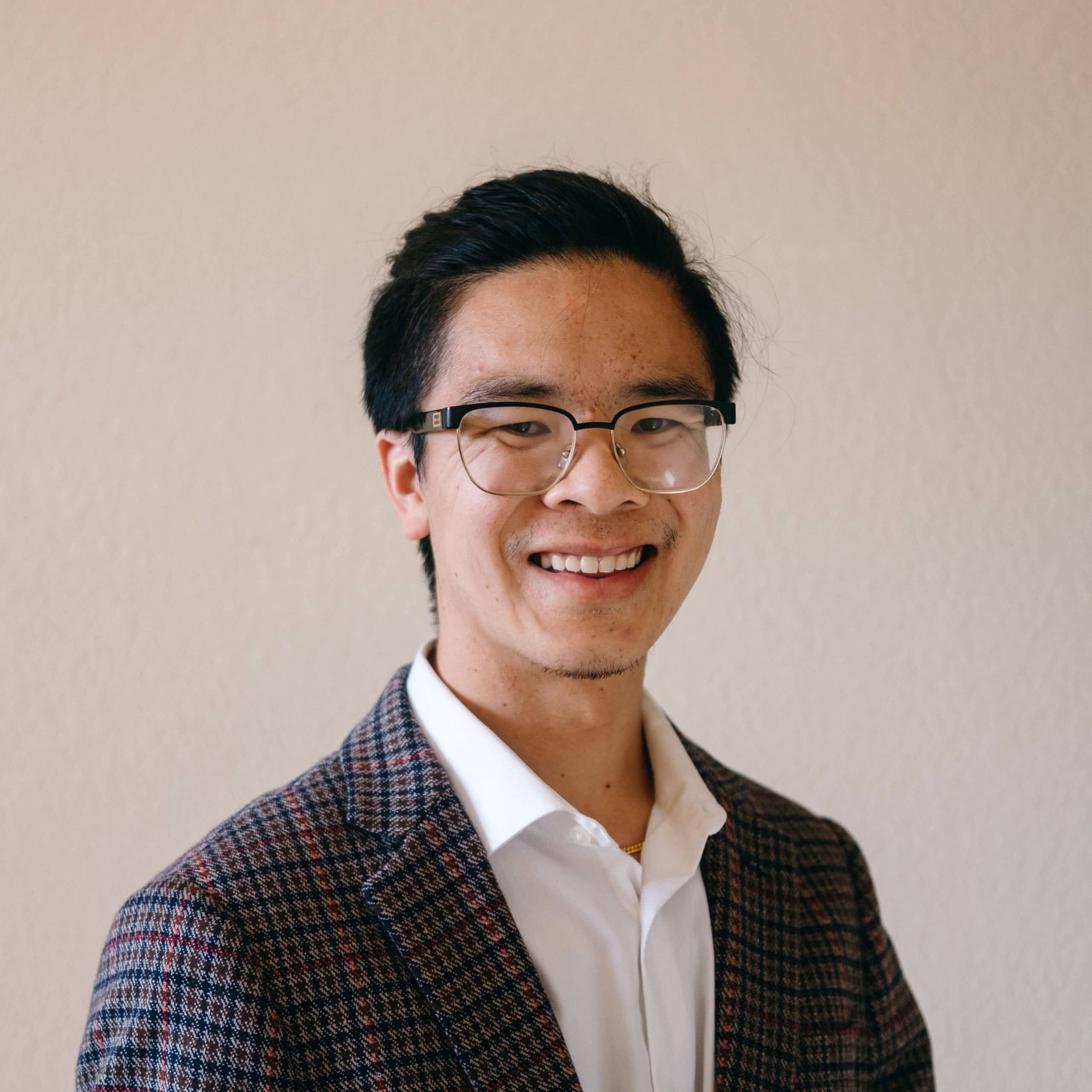Headshot of Curtis Yee. He wears glasses, a white button-up collared shirt, and a plaid overcoat.