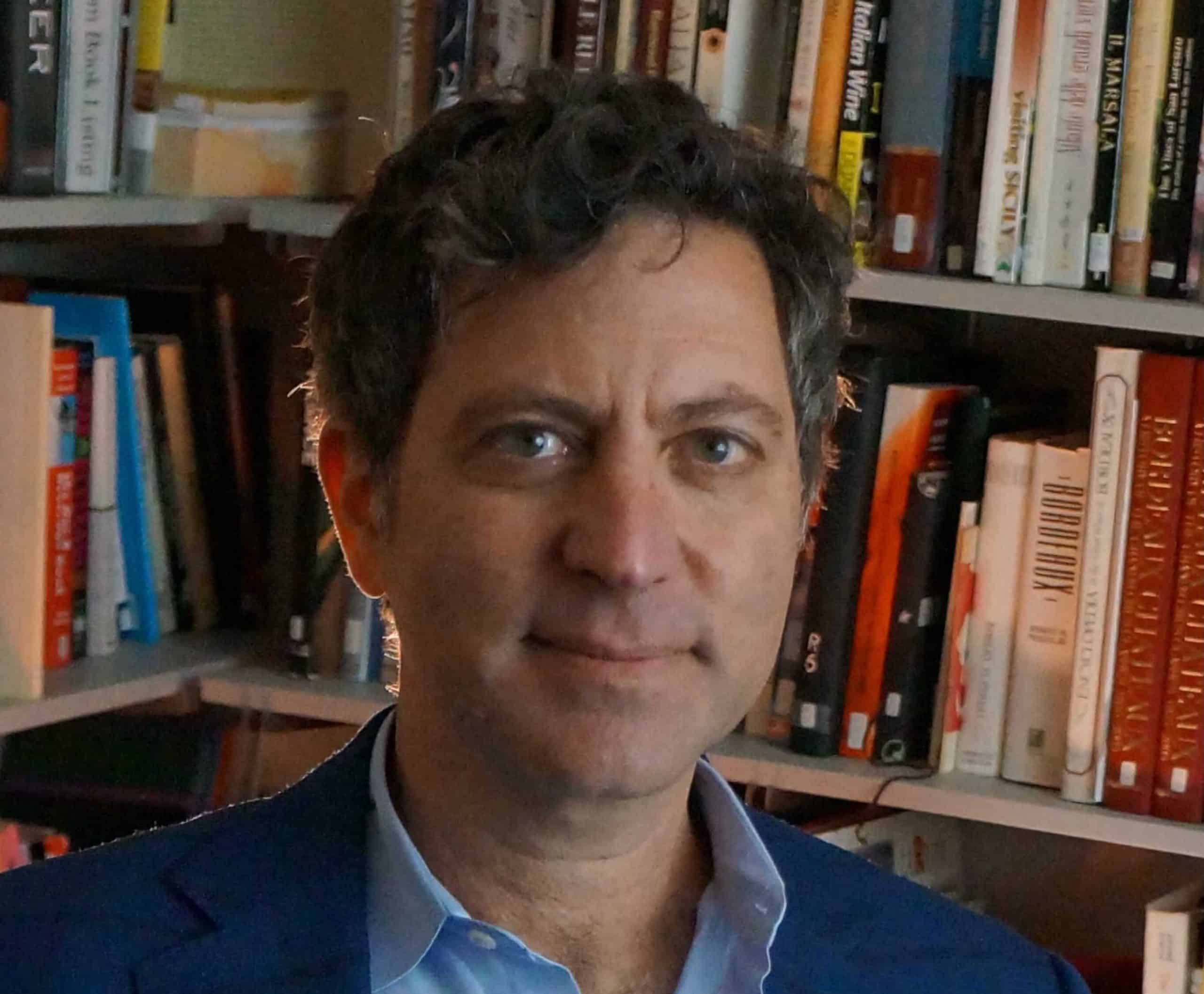 Headshot of Jamal Rayyis. He wear a blazer over a collared shirt and sits in front of a corner bookshelf.