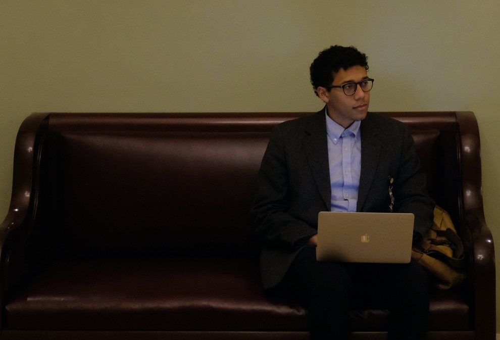 Gabe Schneider sits on a bench. He looks to the right of the frame and is holding a laptop in his lap. He wears glasses, a button up shirt, and a suit jacket.