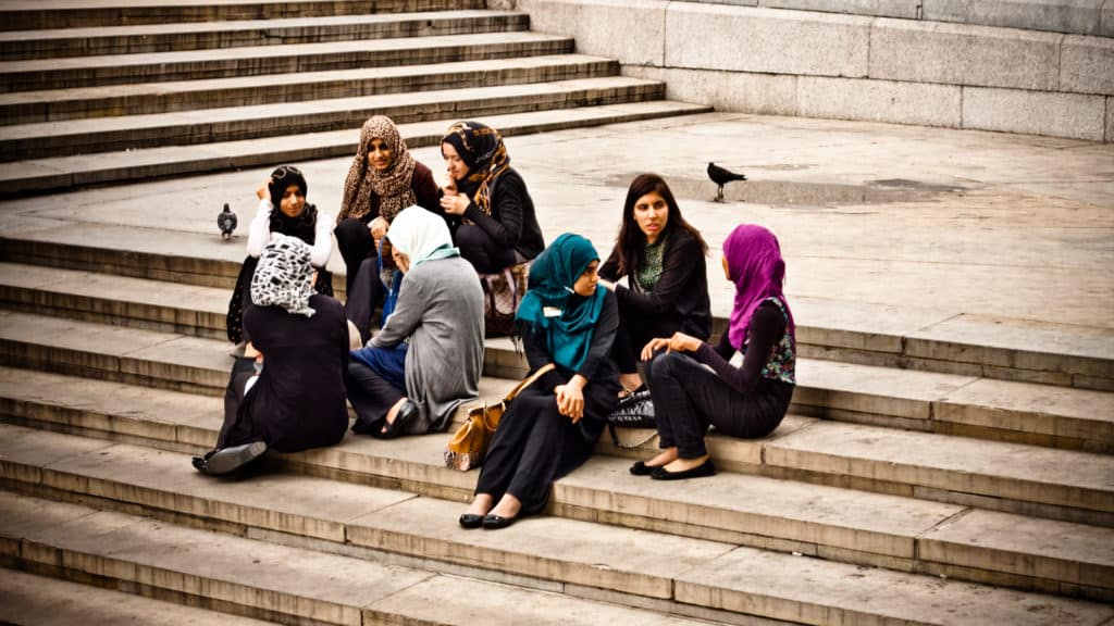 Young Muslim women, some hijabi and others not, sit on the steps of Trafalgar Square talking and laughing.
