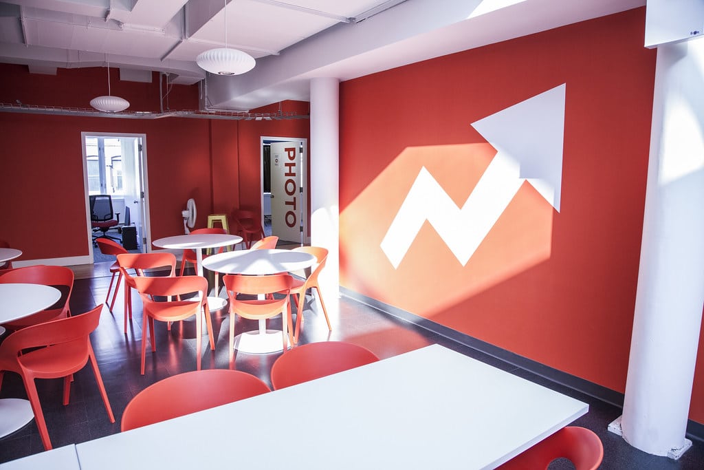 The inside of Buzzfeed's newsroom in New York City. Red walls surround white tables, which sit on a dark gray tile floor. Red chairs circle the tables. The Buzzfeed logo, an upward arrow, is emblazoned and visible on the rightmost wall.