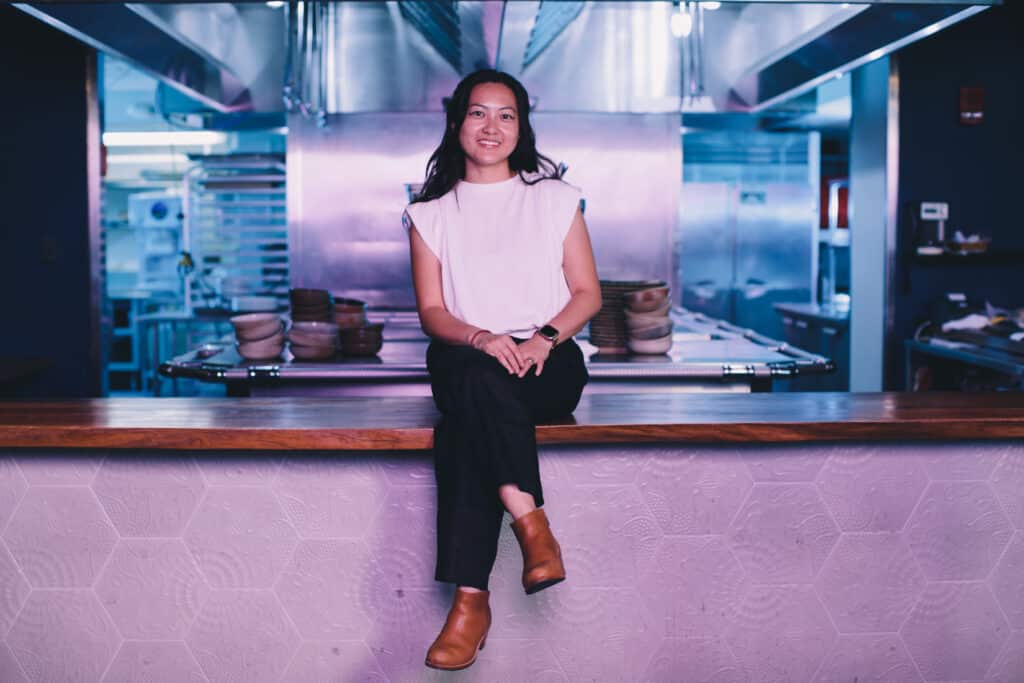 Jenny Dorsey, an Asian woman, sits cross-legged on the counter of an empty kitchen and smiles. 