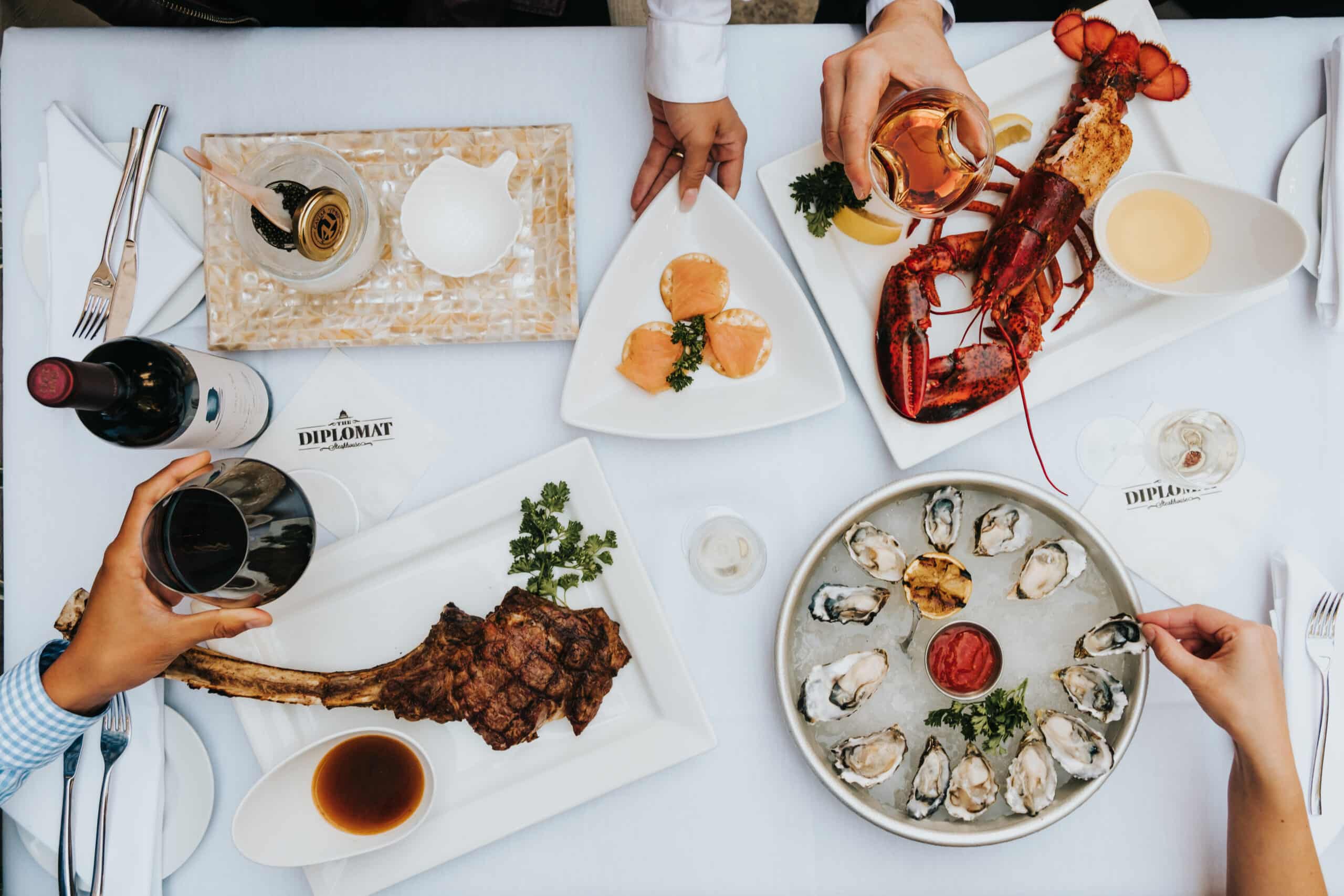 A flat lay of food. Napkins read "The Diplomat," the name of a now-closed Sacramento steakhouse. Among the platters are a dessert, lobster, oysters, and a tomahawk steak. Four diners' hands are visible in the photo. Two diners are holding glasses of wine, red and rosé specifically.