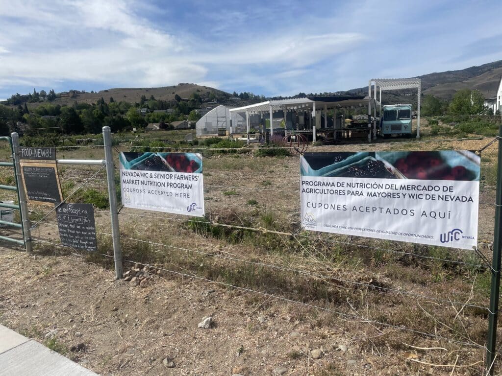 The grounds of Reno Farm Systems. A blue sky with a sheet of clouds hangs over hills dotted with trees and a field with spots of brown and green grass. A barbed wire fence is prominent in front of a greenhouse and other structures. On the fence, white WIC Nevada banners, with blackberry and strawberry baskets, read "Nevada WIC & Senior Farmers' Market Nutrition Program: Coupons Accepted Here" and "Programa de Nutrición del Mercado de Agricultores Para Mayores y WIC de Nevada: Cupones Aceptados Aquí." Square blackboards to the left read "FOOD NEWS!" and have information about plant containers.