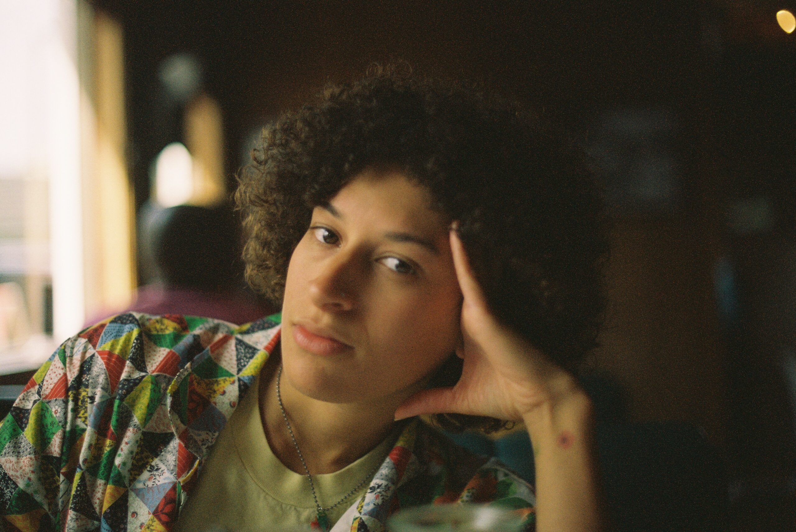 The author, Izzy Johnson, leans their head on their hand, looking at the camera with a neutral expression. They have an afro and are wearing a strawberry necklace, a short-sleeve rainbow patchwork square button-up and a pale green shirt.