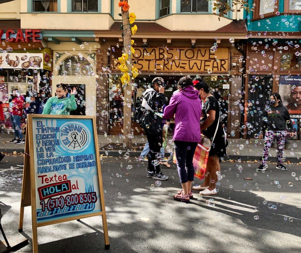 People gather in a street surrounded by a storm of bubbles for a Día de los Muertos celebration next to a Spanish sign from Bay Area news outlet El Timpano, encouraging attendees to sign up for their outlet. The sign reads "El Tímpano escucha y realza la voz de la comunídad Híspano hablante de Oakland. Textea HOLA al 15108008305."
