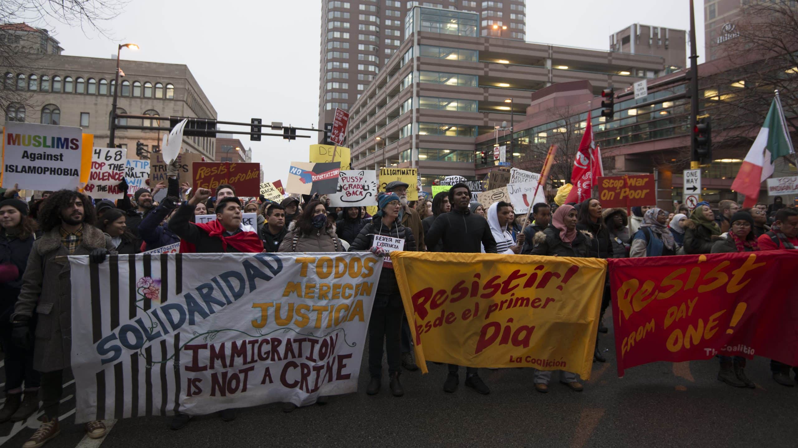 A mass of protesters hold up signs protesting President Trump's stances on marginalized groups. Among the slogans on them are "Muslims Against Islamophobia", "Solidaridad Todos Merecin Justicia — Immigration is not a crime", "Resistir! [D]esde el primer Dia", "Resist from Day One!", "Stop War on Poor", and "You are on Indian Land." 