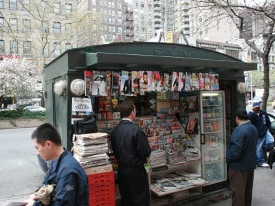 Four people mill around a fully-stocked New York City green newsstand.