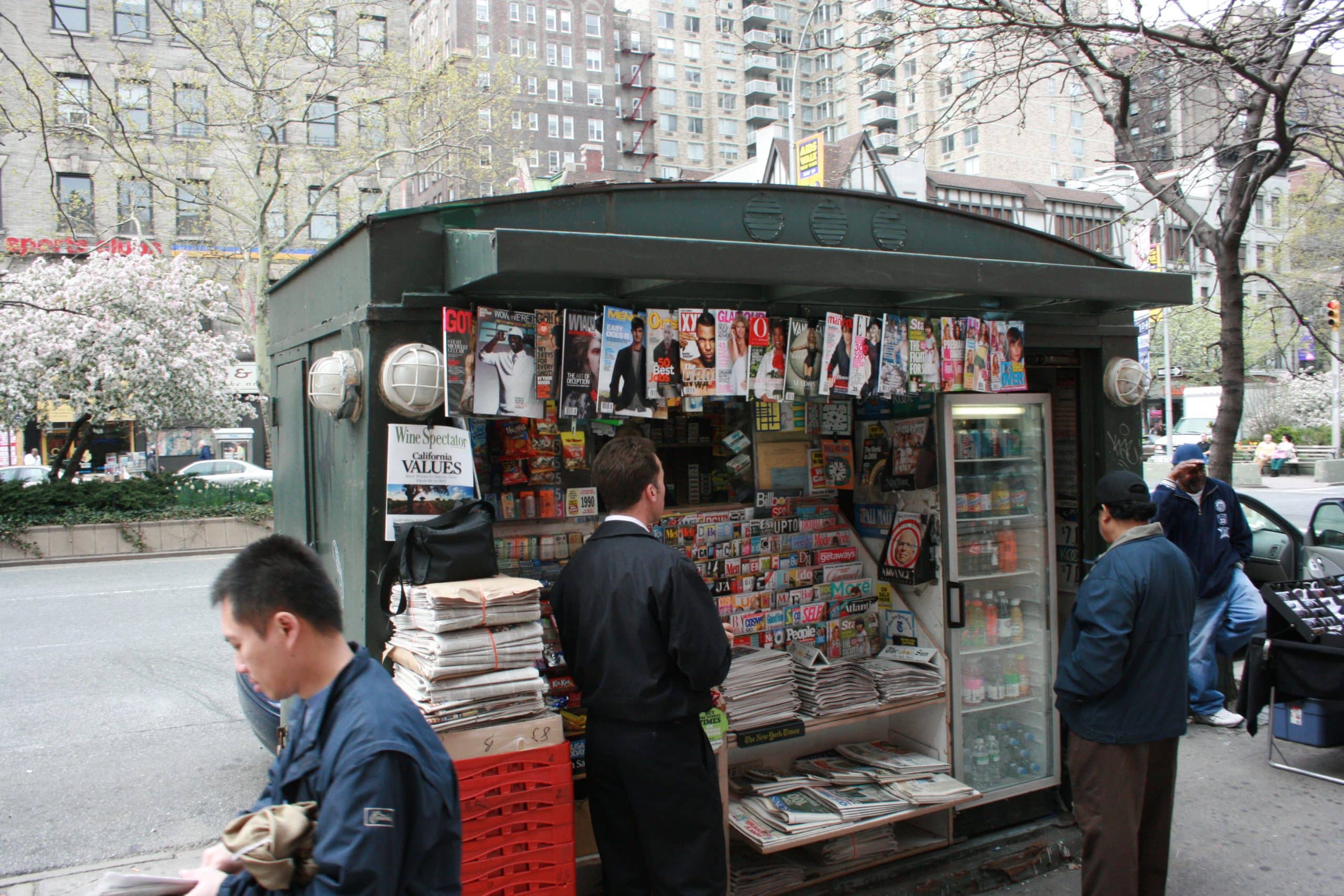 Four people mill around a fully-stocked New York City green newsstand.