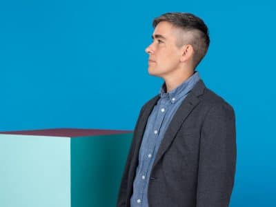 Lewis Raven Wallace, a white trans journalist with a suit, faces left against a blue wall.