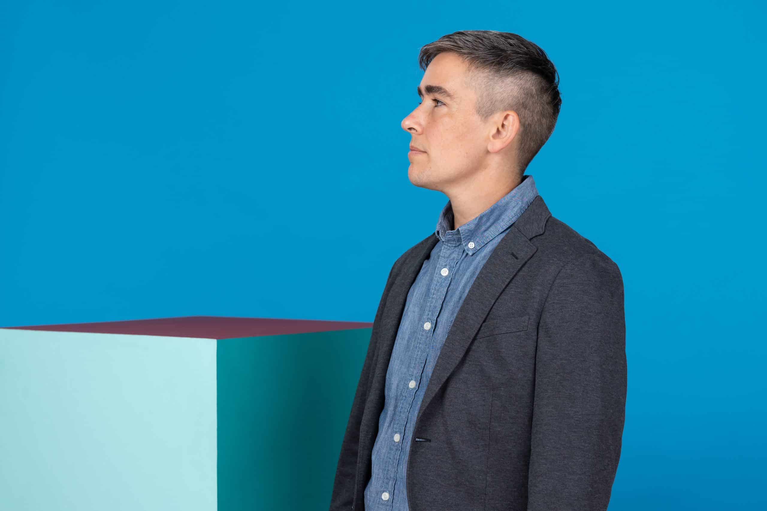 Lewis Raven Wallace, a white trans journalist with a suit, faces left against a blue wall.