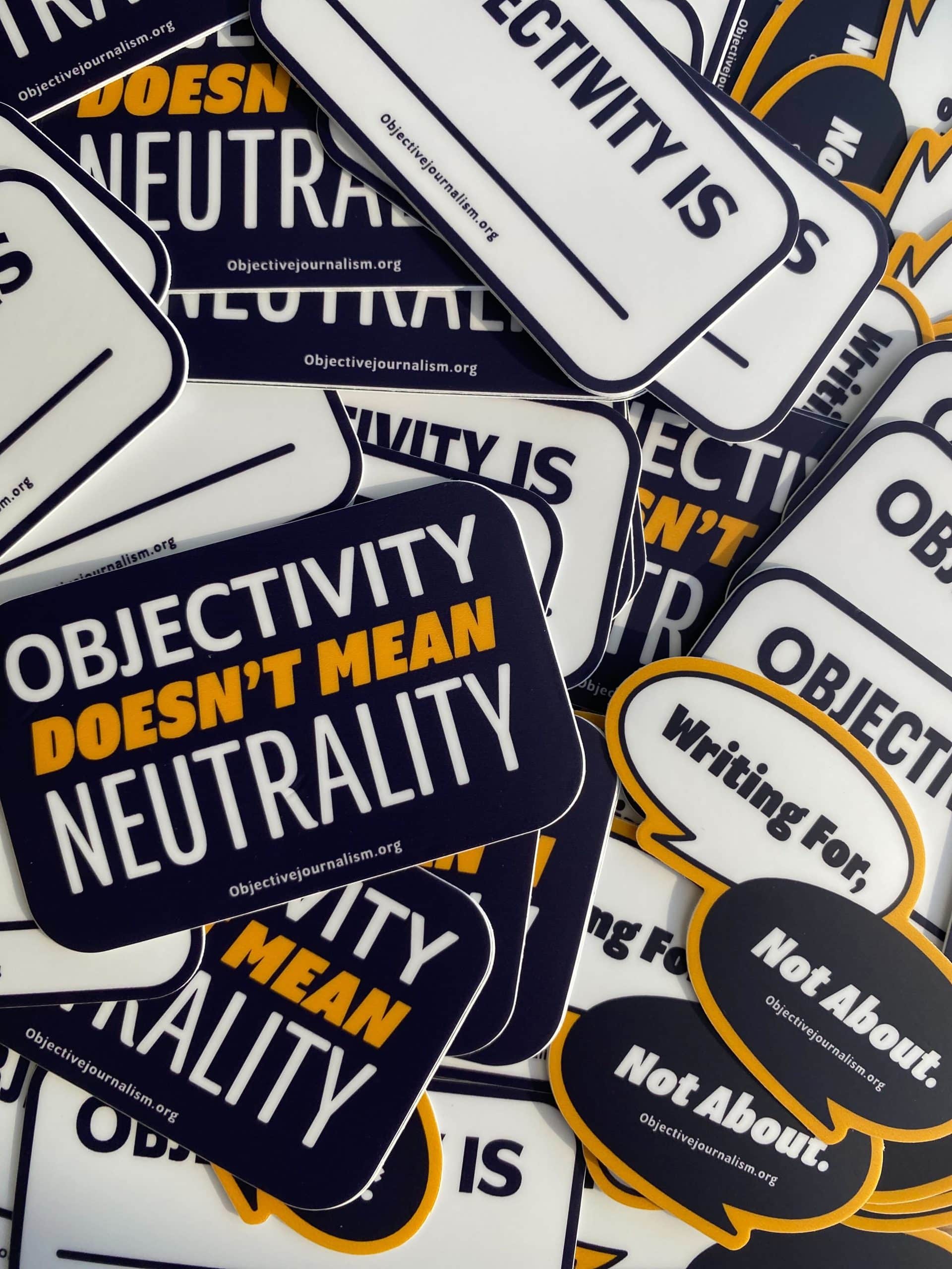 A pile of stickers. The three designs are a fill-in-the-blank sticker that says "Objectivity is [blank]", a navy blue sticker that says "Objectivity doesn't mean neutrality," and a sticker with two speech bubbles — one bubble has the words "Writing for," overlapped by one reading "Not About." 