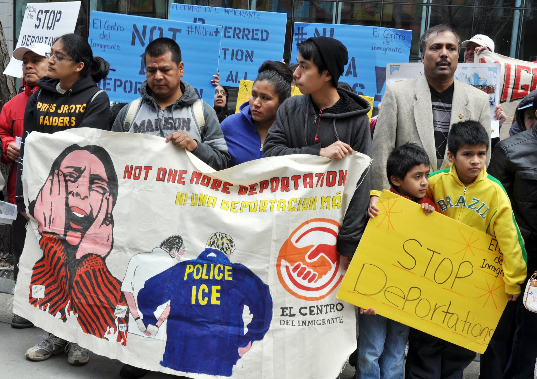 Demonstrators hold up posters protesting deportations. The central banner reads Not One More Deportation, Ni Una Deportación Más. Another prominently displayed poster reads "Stop Deportations."