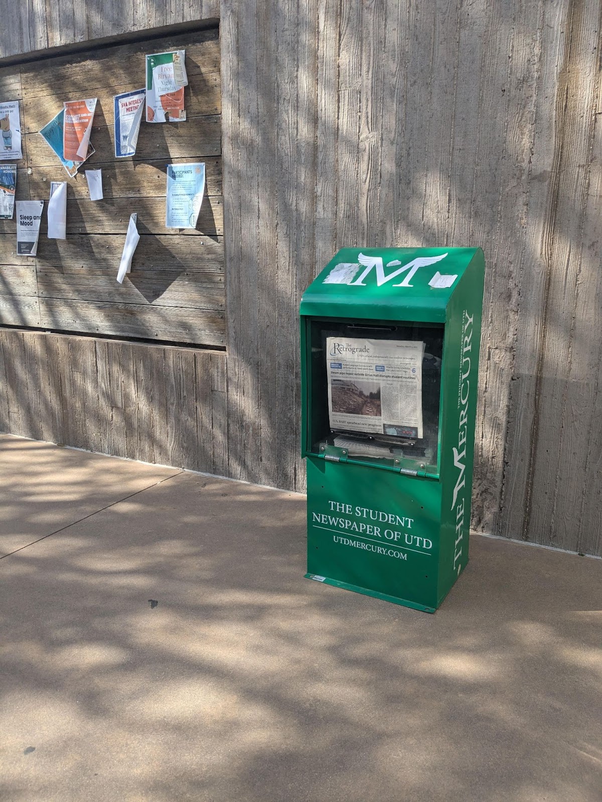 The sole remaining Mercury-branded newsstand sits next to an outdoor bulletin board, filled with issues of The Retrograde.