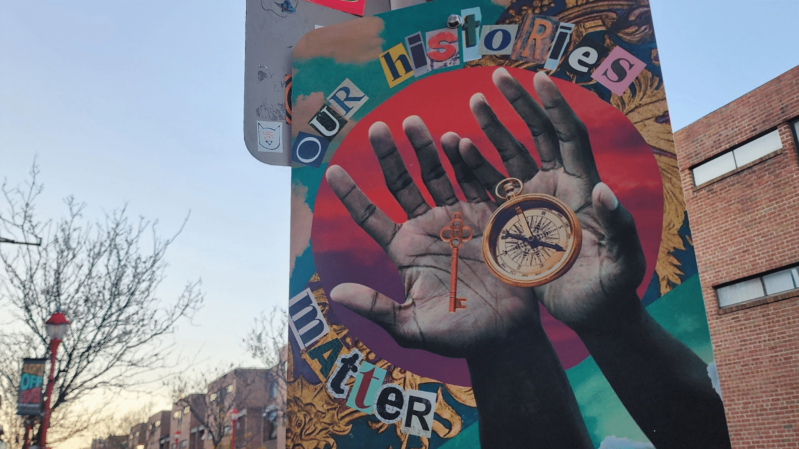 A mural on the back of a lamppost. Cut-out letters read Our Histories Matter around a red and purple circle behind a photo of Black hands holding a key and a compass.