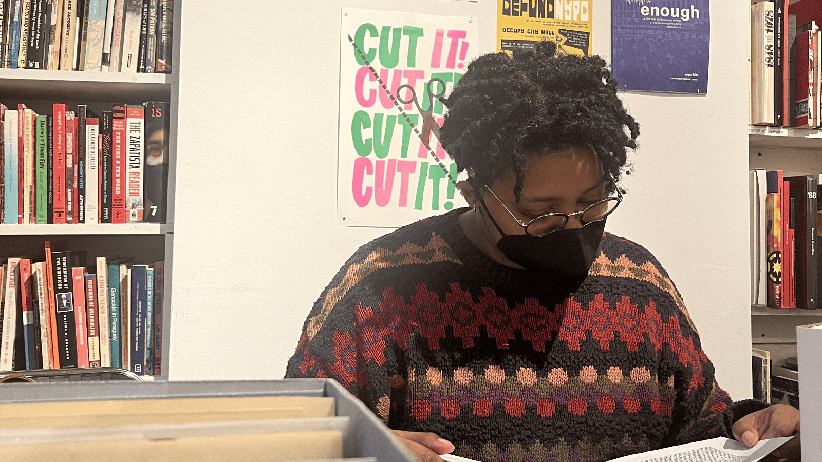 A masked Black person with short twists and glasses pores over records at a library.