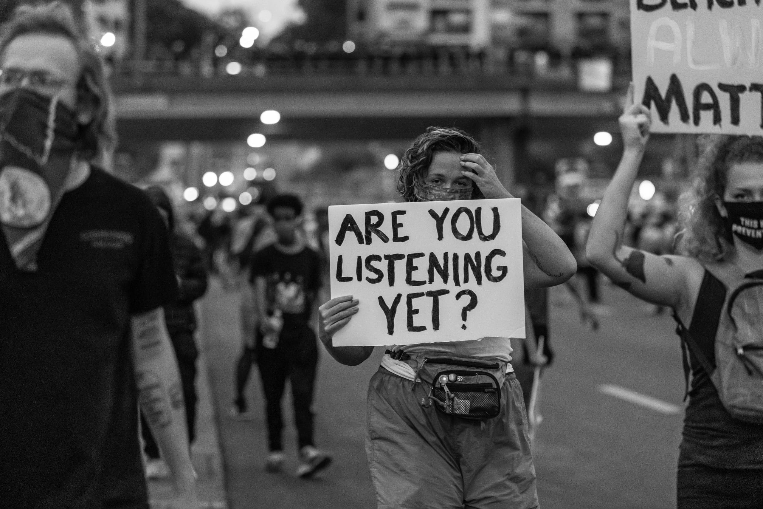 A grayscale photo of a group of masked protesters holding up signs; the foremost says "Are You Listening Yet?"